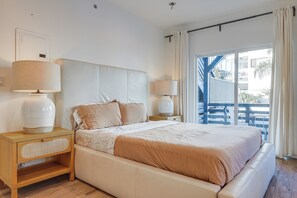 A bedroom with a large bed, two bedside tables with lamps, a wooden nightstand, and a view of a balcony with outdoor furniture. - Sunset Strolls & Seaside Fun: Santa Monica Condo! (Santa Monica)