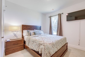 A bedroom with a wooden bed, a nightstand with a lamp, a TV mounted on the wall, and a window with curtains. - Port St. Lucie Home on Florida's Treasure Coast (Port St. Lucie)