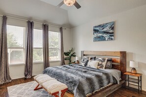 A bedroom with a large bed, a bedside table with a lamp, a window with curtains, and a painting on the wall. - Home Theater & Game Room! 7BR San Antonio Retreat (San Antonio)