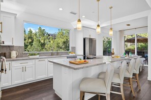 A modern kitchen with a large island, white cabinetry, and a view of a forest. - Covered Lounge + Fire Pit + BBQ | Red Rock Views + Mins to Town | Casa Alta (Sedona)