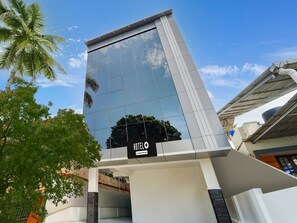 A modern hotel building with a glass facade, a sign indicating it's a company serviced hotel, and a palm tree in the foreground. - Saver Double Pullans Arcade (Kanayannur)