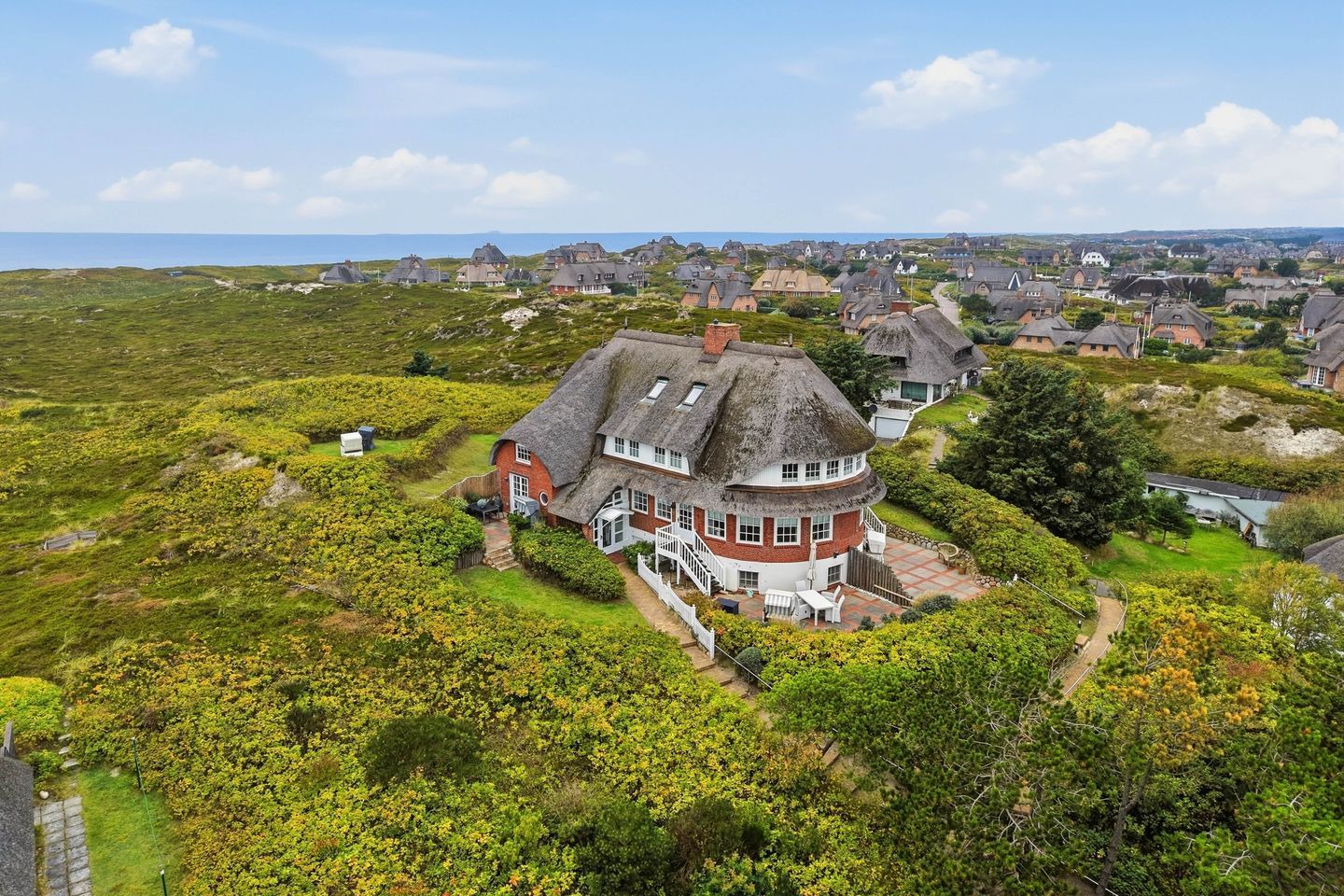 A large house with a circular layout, surrounded by lush greenery and a coastal view.