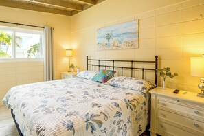 A bedroom with a bed, a nightstand, a lamp, a painting on the wall, and a window with a view of palm trees. - 1/2 Mi to Ocean! Canal-Front Key Colony Beach Home (Key Colony Beach)