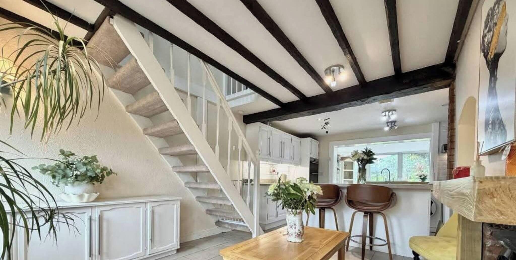 A staircase leading to a kitchen with white cabinets, a wooden table with a vase, and two bar stools.