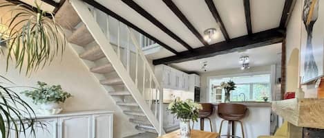A staircase leading to a kitchen with white cabinets, a wooden table with a vase, and two bar stools.