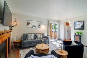 Ein Wohnzimmer mit einer Couch, zwei runden Tischen, einem Flachbildfernseher, einem Kamin und Blick durch eine Glastür nach draußen.