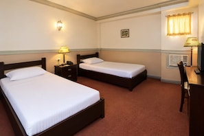 Quarto de hotel com duas camas de solteiro, uma escrivaninha com cadeira, luminária, telefone, quadro na parede e janela com cortinas.