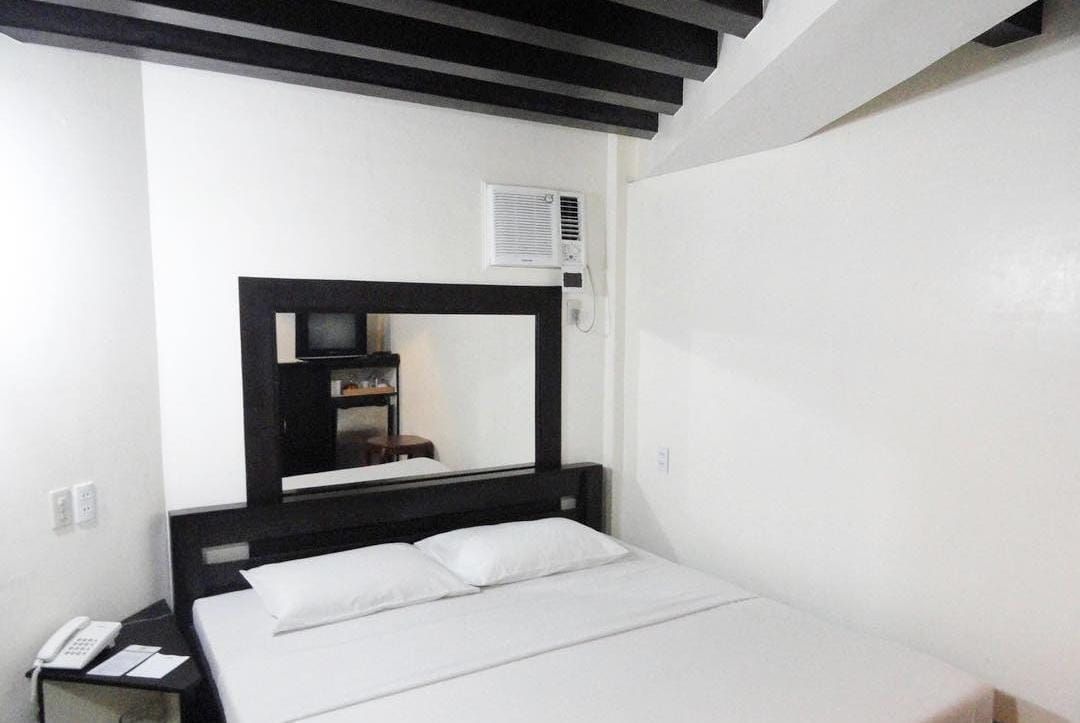 Una habitación de hotel con una cama, una mesita de noche con un teléfono, un espejo y un equipo de aire acondicionado de pared.