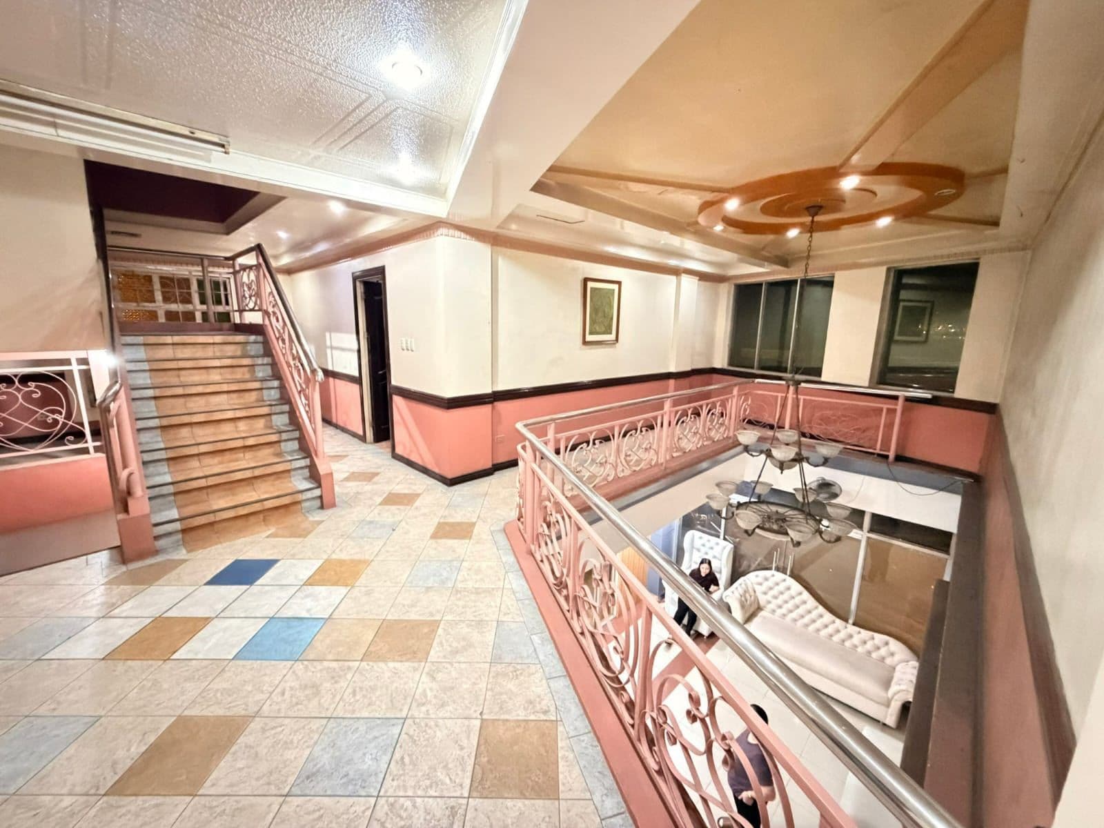 Een hotellobby met een trap, een zithoek met een bank en een tafel, en een plafondventilator.