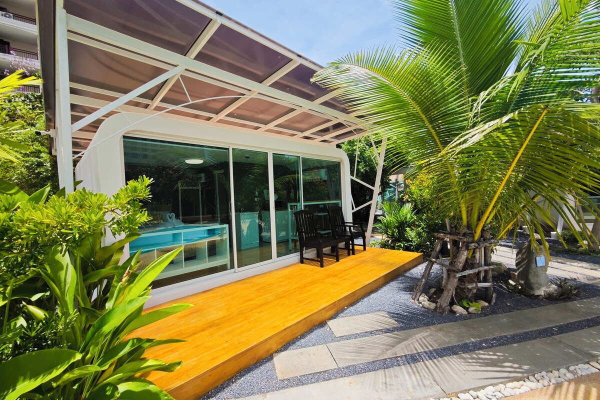 Un área de piscina moderna al aire libre con terraza de madera, paredes de cristal y abundante vegetación.
