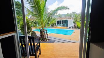 A view of a swimming pool with a wooden deck and a palm tree, a covered seating area, and a building in the background.