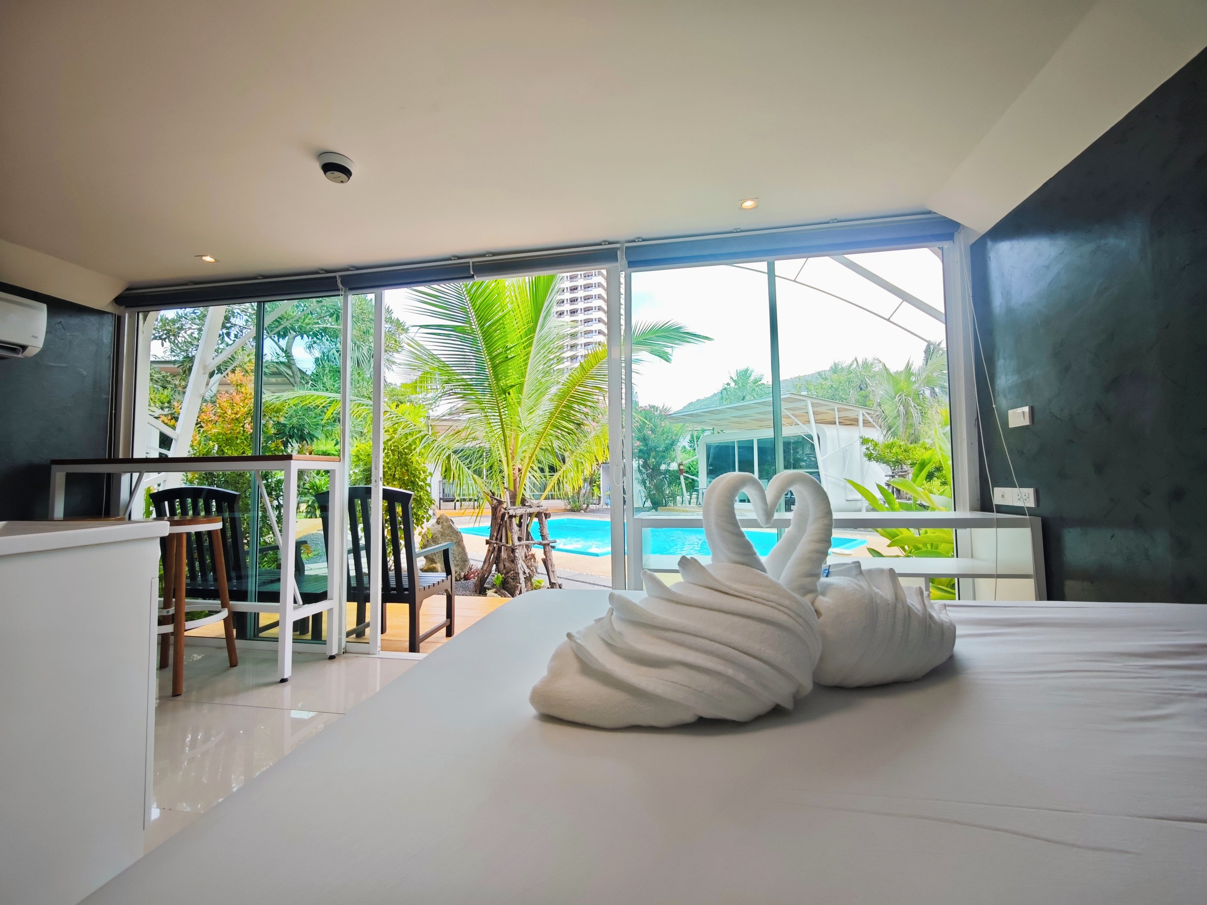 A modern hotel room with a large bed, a swan-shaped pillow, a balcony with a view of a swimming pool and palm trees.