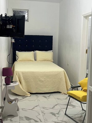Ein Schlafzimmer mit einem Bett, einem Nachttisch mit Lampe, einem Sessel und einem an der Wand befestigten Fernseher.