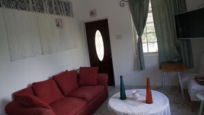 Un salon avec un canapé rouge, une table ronde ornée de vases, une porte avec un vitrail et un téléviseur posé sur un meuble.