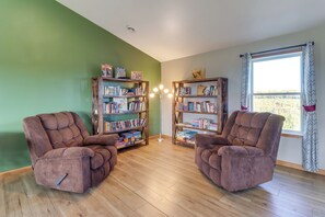 A room with two brown armchairs, two wooden bookshelves filled with books, a window with patterned curtains, and a wooden floor.