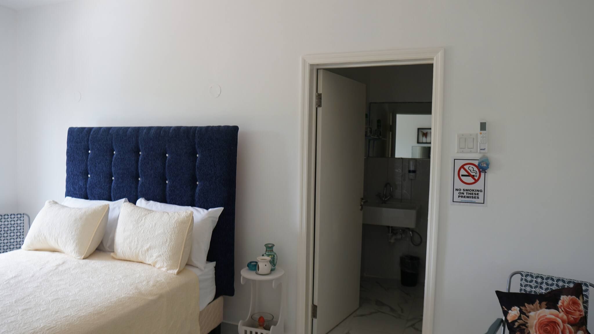 Een slaapkamer met een bed, nachtkastje, deur naar een badkamer en een rookvrij sign op de muur.