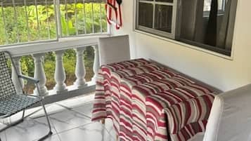 Quarto com cama, uma cadeira, janela com cortinas e vista para o verde lá fora.