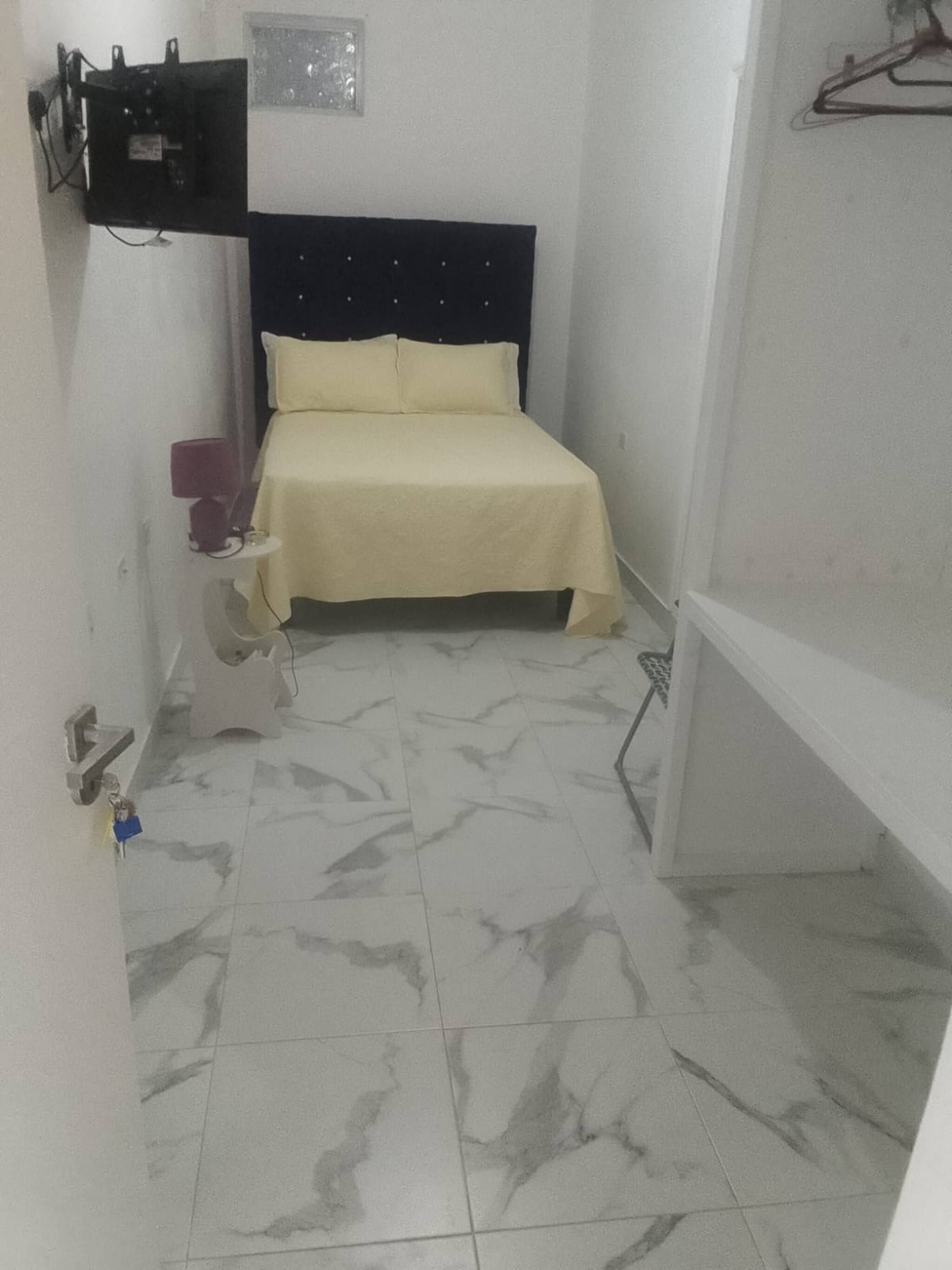 A small, narrow room with a bed, a wall-mounted TV, and a tiled floor.