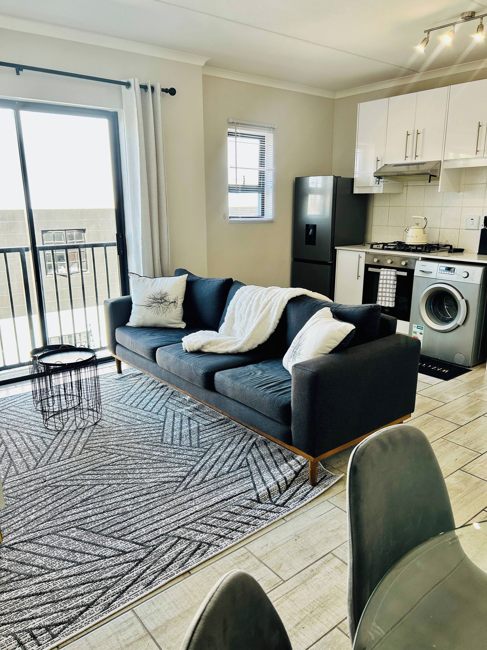 A modern living room with a sofa, a dining area with chairs, a kitchen with appliances, and a balcony with a railing.