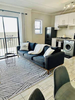A modern living room with a sofa, a dining area with chairs, a kitchen with appliances, and a balcony with a railing.