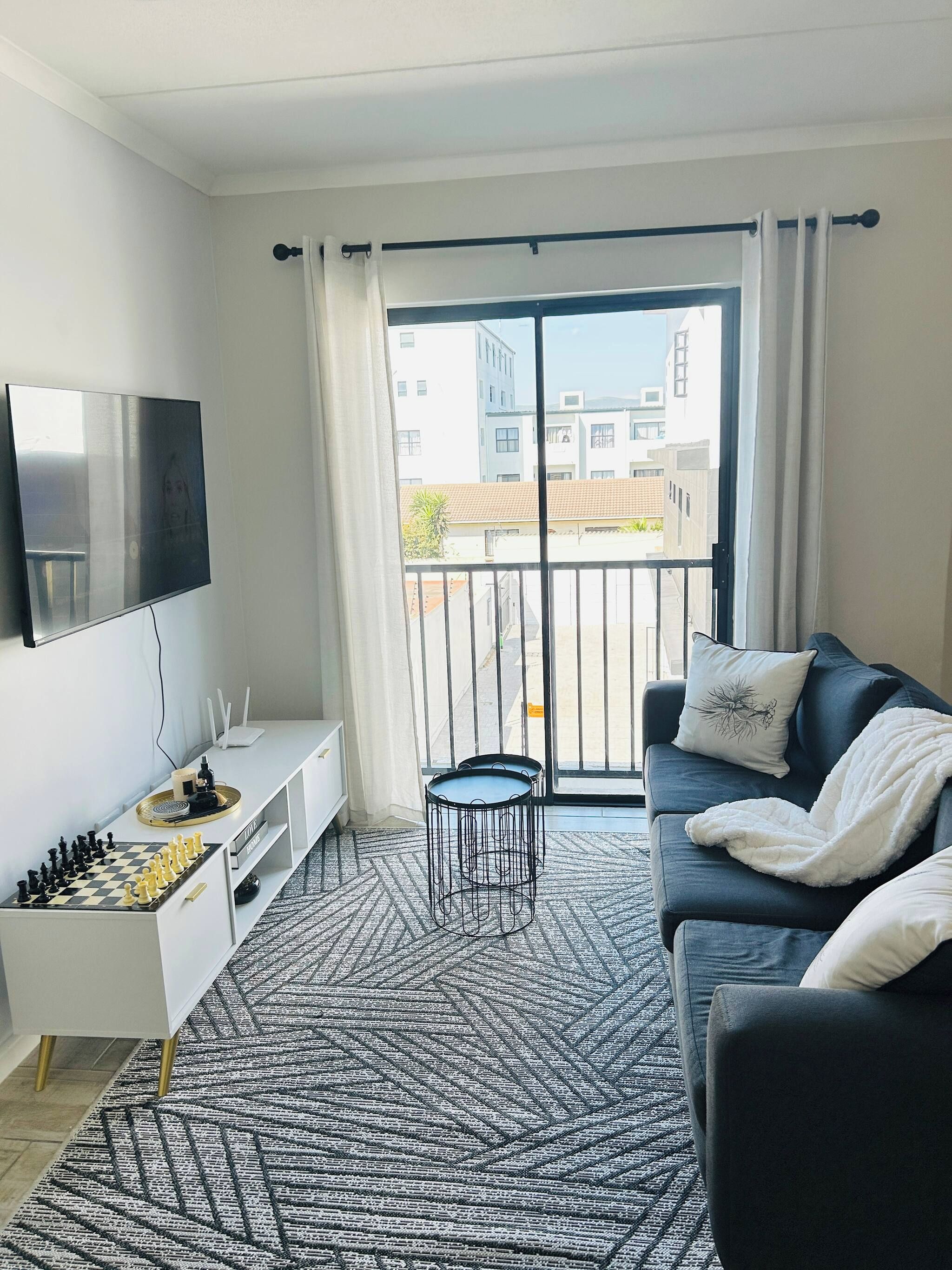 A modern living room with a flat-screen TV, a chessboard, a sofa with cushions, a small side table, and a patterned rug.