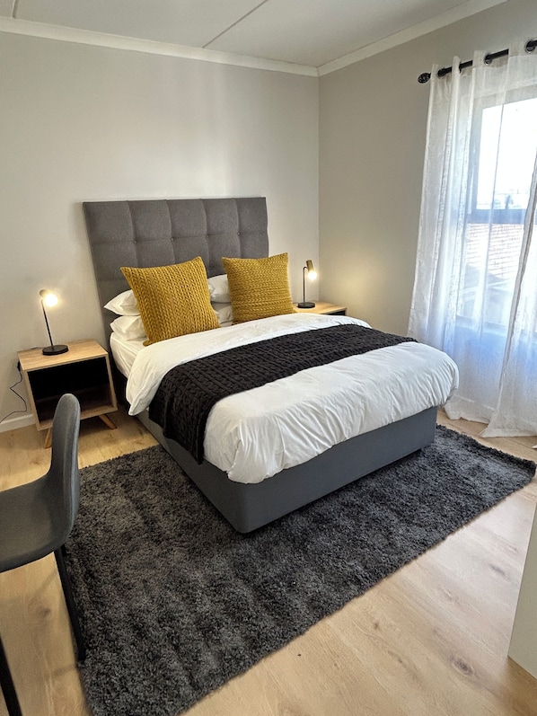 A modern bedroom with a grey upholstered headboard, a black and white bedspread, two mustard-colored pillows, a black throw blanket, a grey chair, and a small bedside table with a lamp.