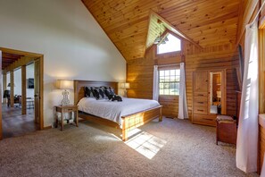 Une chambre avec un plafond en bois, un lit recouvert de linge de lit blanc, une table de chevet avec une lampe, et une vue sur l’extérieur par une fenêtre.