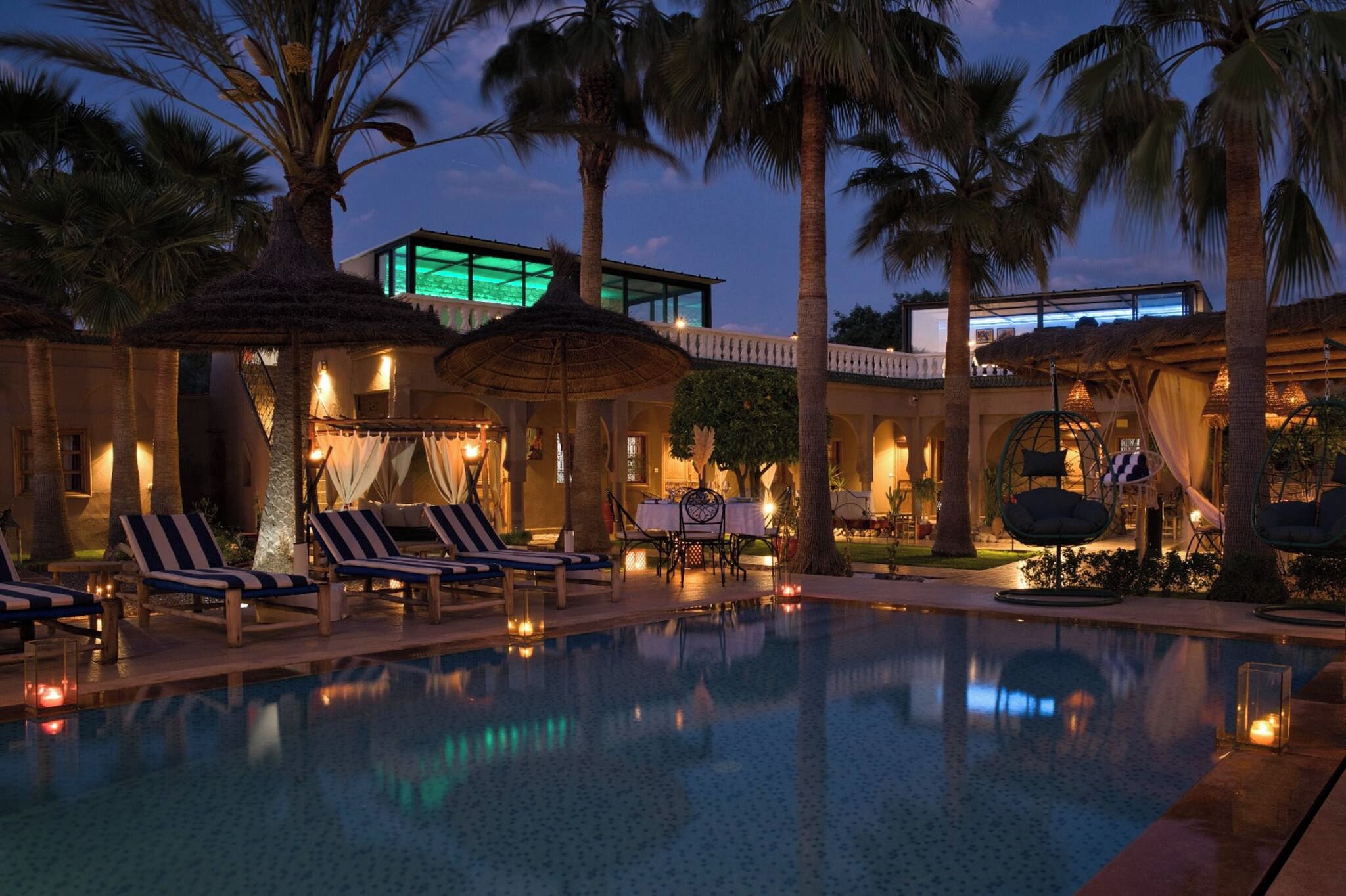 Un espace piscine avec des chaises longues, des parasols et un bâtiment dont une fenêtre est illuminée de lumière verte.