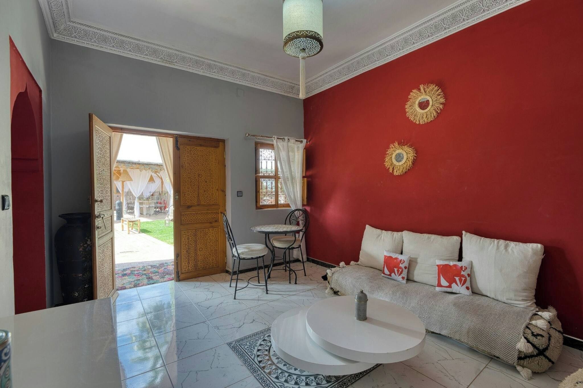 Une pièce avec un mur rouge, un canapé blanc agrémenté de coussins décoratifs, une table basse ronde, un tapis à motifs et une porte donnant sur un espace extérieur.
