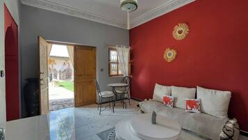 Une pièce avec un mur rouge, un canapé blanc agrémenté de coussins décoratifs, une table basse ronde, un tapis à motifs et une porte donnant sur un espace extérieur.