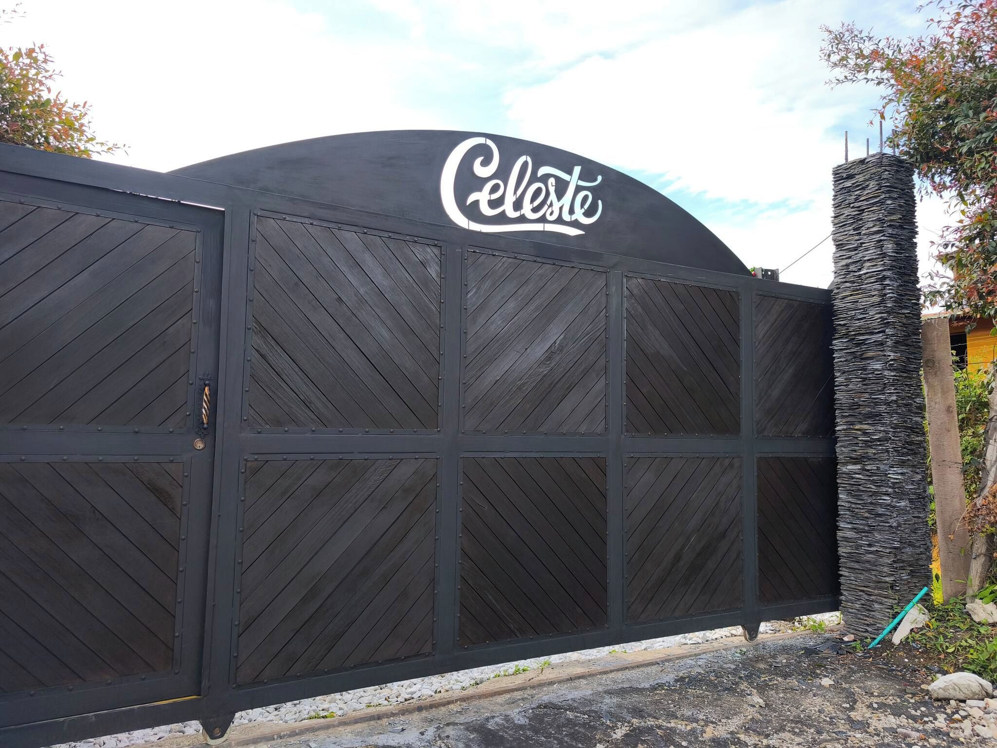 En svart metallgrind med en skylt med texten ”Celeste” ovanpå, en stenkantare till höger och en grusgång framför.