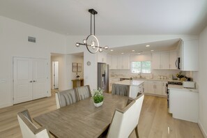 A modern kitchen with white cabinets, a central island, and a dining area with a wooden table and chairs. - Coachella Getaway w/ Yard! Near Festivals & Golf (Coachella)
