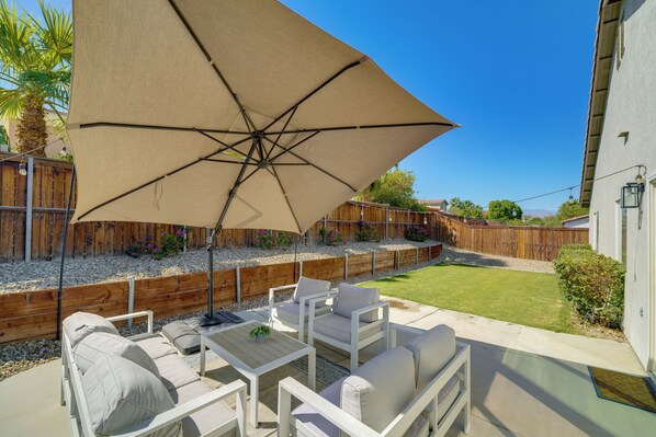 Un patio al aire libre con un paraguas beige, mobiliario blanco y una cerca de madera.