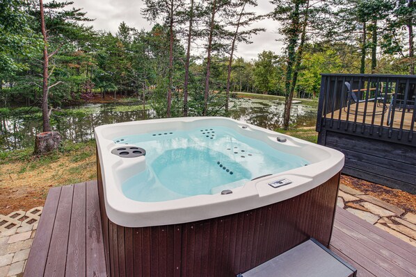 A hot tub on a wooden deck with a pond and trees in the background.