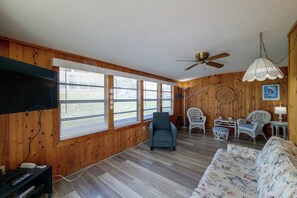 A room with wooden walls, a ceiling fan, a flat-screen TV, a blue armchair, wicker chairs, a floral sofa, and a small table with decorative items. - The Wandering Parrot: Pool, Pickleball & Lake (Clermont)