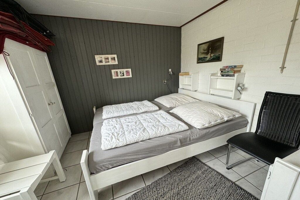 A single-bed room with a grey wall, a white door, a bedside table, a chair, and a small shelf with books.