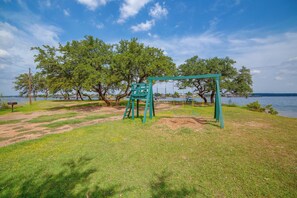 A grassy area with a swing set and a bench near a body of water. - Community Boat Ramp! Family Escape w/ Fenced Yard (Granbury)