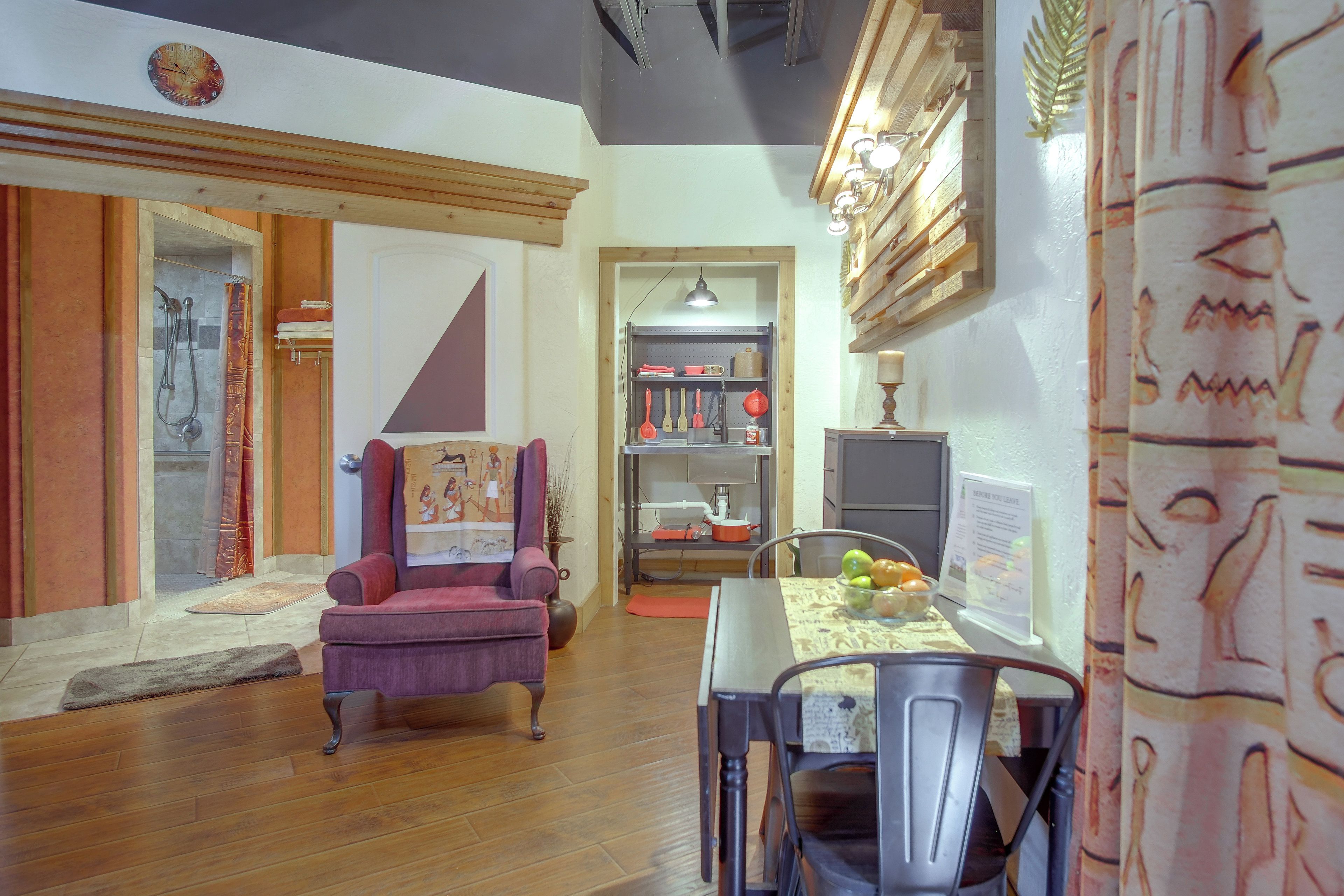 Una habitación con piso de madera, un sillón morado, una mesa de comedor con sillas, un televisor montado en la pared y un estante con utensilios de cocina.