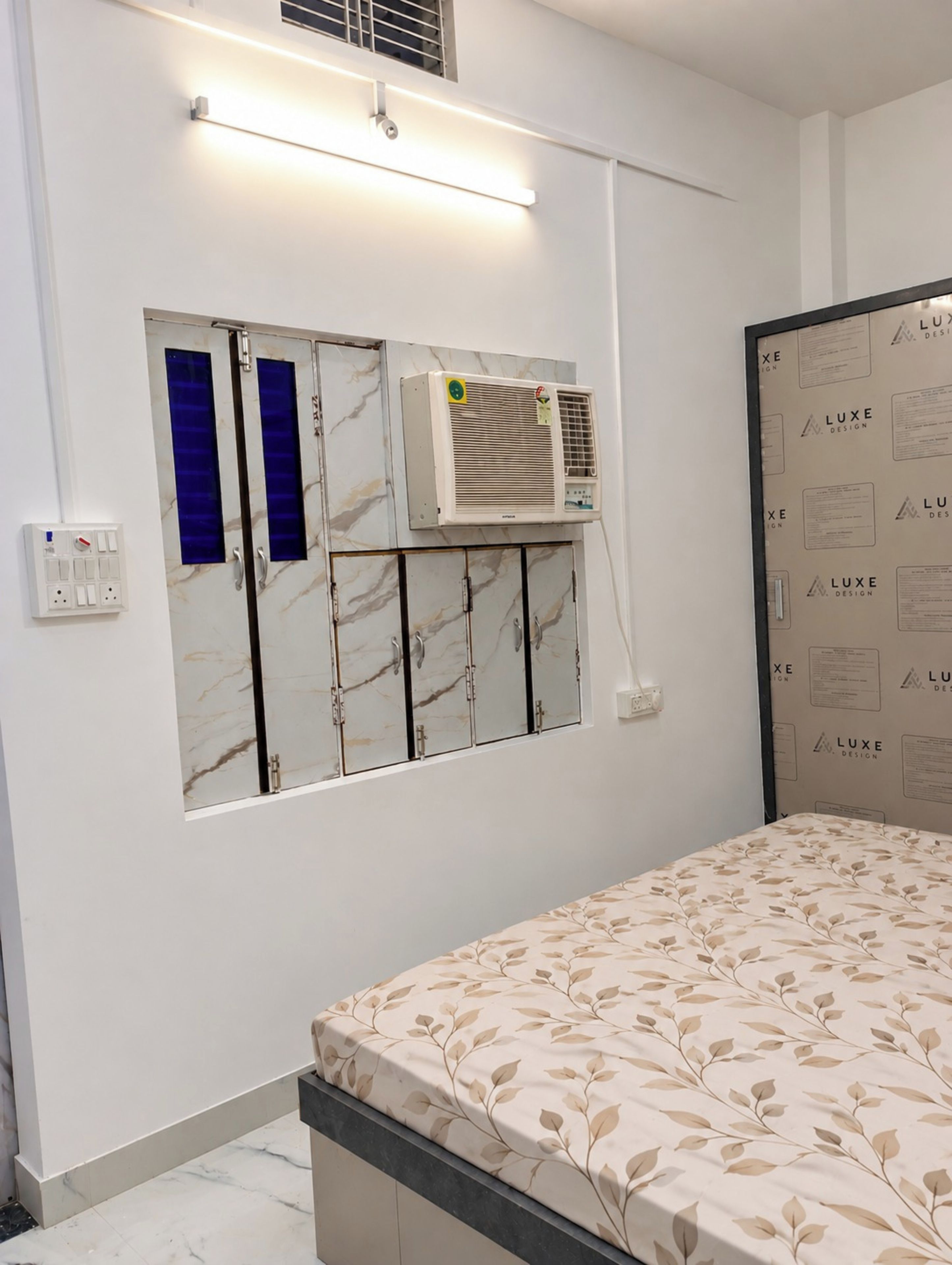 A room with a bed, an air conditioning unit, a wall-mounted cabinet with blue doors, and a wall with printed text.