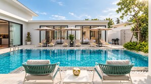 A swimming pool with lounge chairs and umbrellas, surrounded by a modern building with large windows. - Spacious 5-bedroom Private Pool Villa, 10 Min to Bang Tao Beach (Tambon Choeng Thale)