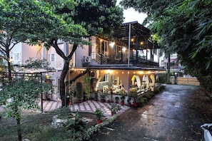 Un edificio de dos pisos con un área de estar cubierta al aire libre, rodeado de follaje.