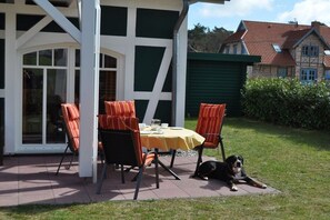Eine Terrasse im Freien mit Tisch und Stühlen, ein Hund liegt im Gras, und im Hintergrund ein Gebäude mit grüner Fassade.