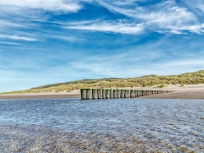 A beach with a wooden structure extending into the water, a sandy shore, and a grassy hill in the background. - "t Hof van Schouwen (Burgh-Haamstede)