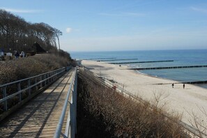 A coastal walkway with a metal railing, leading to a sandy beach with a pier extending into the water. - Bungalow with 2 bedrooms (Ostseebad Nienhagen)