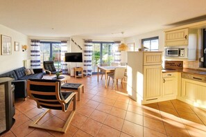 A modern living room with a sofa, a wooden chair, a dining table with chairs, a flat-screen TV, and a kitchen area with cabinets and a microwave. - Holiday home in Zingst with swimming pool (Zingst)