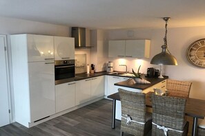 A modern kitchen with white cabinets, a stainless steel range hood, and a dining area with wicker chairs and a wooden table. - Holiday apartment with 2 bedrooms (Grömitz)