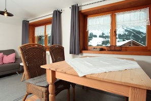 A wooden dining table with a white tablecloth, two wicker chairs with grey cushions, a grey sofa with a red and white checkered pillow, and a window with a view of snow-covered mountains. - Holiday apartment with 2 bedrooms (Riezlern)
