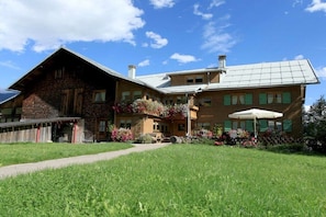 A two-story building with a brick facade, a covered porch, and a garden with flowers. - Holiday apartment with 2 bedrooms (Riezlern)