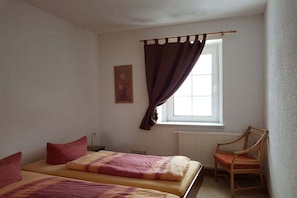 Quarto com duas camas, uma cadeira, janela com cortinas e radiador.