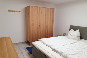 A bedroom with a bed, a wooden wardrobe, a nightstand, and a blue rug. - Holiday apartment with terrace and view (Petersdorf\/ Fehmarn)
